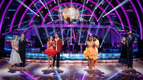 BBC/PA The four remaining couples line up on the ballroom floor. Karen Carney is wearing a blue dress. George Clarke and his partner Alexis Warr wear red. Balvinder Sopal is in an orange dress and Amber Davies wears a dark ballgown.