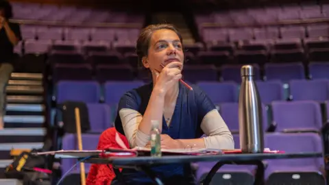 Lillie Sherry Rosie sits in the auditorium at a desk with pencil in hand surrounded by purple chairs.