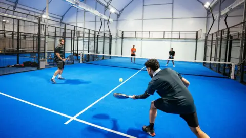 Four people play a match on a blue padel court. Two players in the foreground are positioned close to the camera with one preparing to hit a ball with a racket. The court is enclosed by metal fencing and is inside a large, high‑roofed structure.