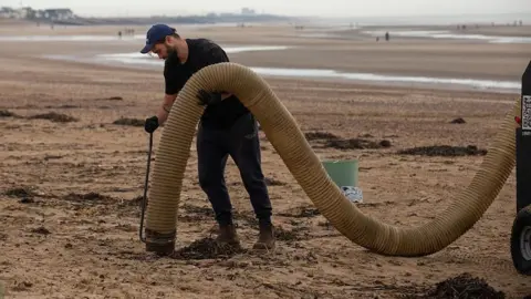 A man is on a beach, holding a large plastic hose. He's dressed in black, with a navy blue cap