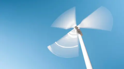 Getty Images A view from the ground of a white wind turbine, its three blades blurred as they spin against a clear blue sky.