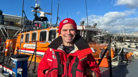 A man in a red coat and red beanie smiles to the camera. He stands in front of an orange lifeboat called Sir Max Aitken III. 