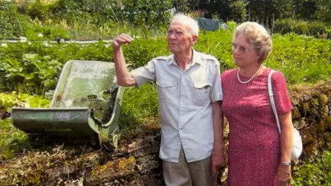 An older man and woman standing next to a grass verge. On the verge is a green piece of metal, which is the shell of a seat.