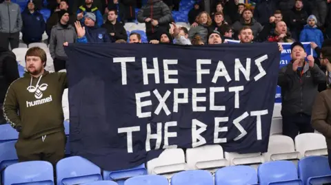 Fans hold a bedsheet banner saying 'The Fans Expect The Best' in a football stadium.