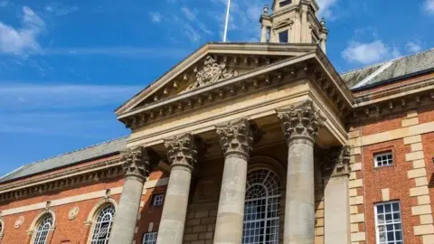 BBC A red brick building with large windows. In the front are four concrete pillars with a triangular roof above them.