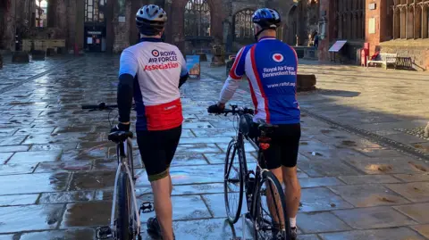 Stuart Green Stuart and Roger are with their bikes again, this time we are seeing them from behind. They are to the side of their bikes, looking ready to climb back on and get going.