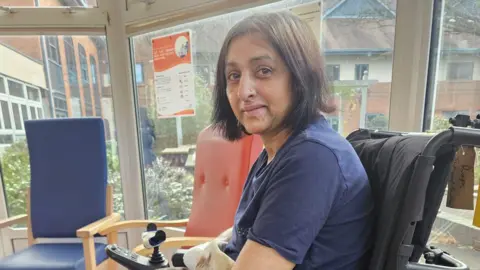 Kam Sangha Manjit is seated in a powered wheelchair near large windows. She is smiling. Chairs in different colours are placed around the room, suggesting a communal or rehabilitation setting.

