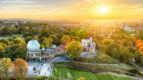 Getty Images The Royal Observatory as seen from the sky. It has a large domed building on one side and a plaza at the front. In the background is the London skyline and the setting sun.