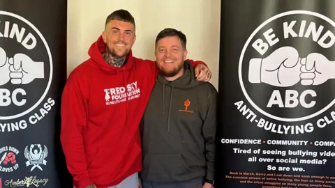 Troy Baker, left, has his arm around Gerry Ferguson's shoulder as they smile at the camera. Mr Baker is wearing a red hoodie with Red Sky Foundation written across it in white. Mr Ferguson is wearing a grey hoodie with an orange logo. They are standing between two black banners reading Be Kind ABC ANTI-BULLYING CLASS. the logo is of two fists, a larger one and a smaller one, presumably of a child, bumping into each other. 