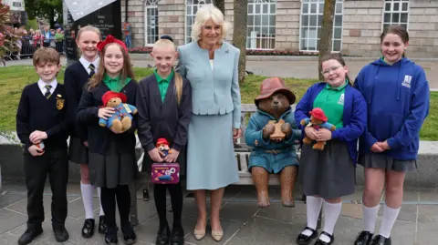 Reuters Britain's Queen Camilla meets children during her visit to Conway Square, in Newtownards, Northern Ireland. They are standing in front of a statue of Paddington Bear which is sitting on a bench. The Queen is wearing a light blue skirt suit. Four schoolchildren are standing to her right and two on her left. They are five girls and one boy and three of the girls are holding Paddington Bear toys.