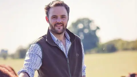 James Florey James Florey is wearing a shirt under a brown gilet and is touching a cow. He is standing outside in a field.