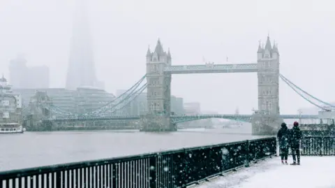 Getti images of Tems and Tower Bridge River in London on an icy day. In the lower right corner, two people are wearing black coats and hats that see the view behind the black railing.