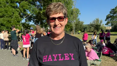 Cameron Noble/BBC A head and shoulders image of Hayley Judge. She is wearing a black T-shirt with the word Hayley written on it in pink. She is wearing dark sunglasses and looking directly into the camera. Other Ed Sheeran fans can be seen in the background.