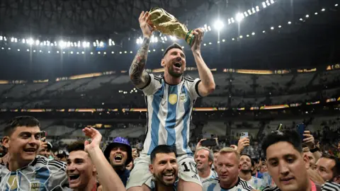 Lionel Messi lifts the World Cup aloft whilst being carried on the shoulder of countryman Sergio Aguero inside a football stadium. They are surrounded by others celebrating Argentina's win and filming on their mobile phones.