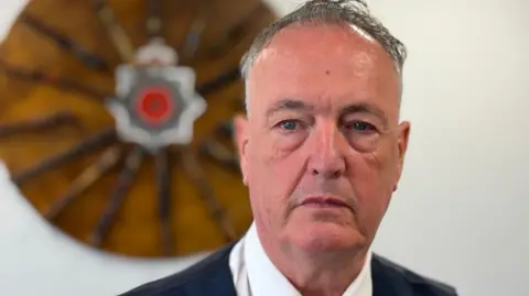 Clive Grunshaw, who is wearing a white shirt, waistcoat and tie, is taring directly at the camera. He is standing in front of the Lancashire Police logo.