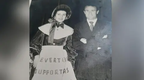 Everton FC toffee lady Mary Morgan (nee Gorry) wearing Victorian style confectionary attire with white pinny and bonnet with Tommy Eglington who played for Everton wearing a suit taken between 1953 and 1956.  