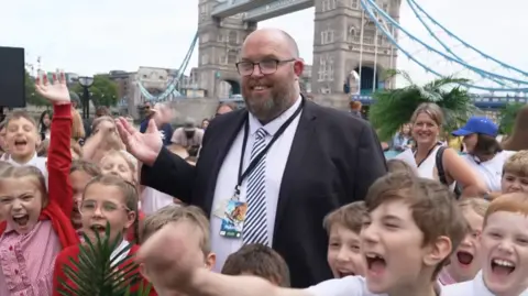 Joshua Falcon/BBC A man wearing a black suit blazer over a white shirt with a striped tie stood in the middle of a group of schoolchildren who are wearing white and red uniforms. In the background is Tower Bridge.
