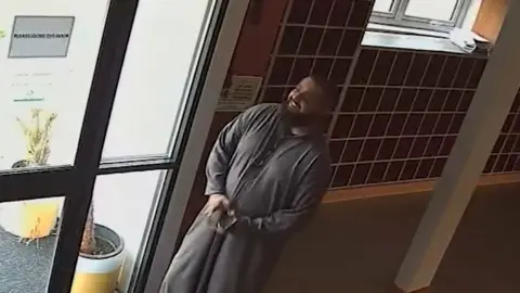 CCTV view of a man in a dark grey outfit smiling by the door of a hallway. Another man sits in the corner putting on a pair of shoes.