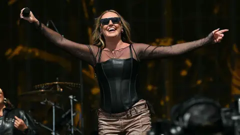 Getty Images Perrie performs on stage at the TRNSMT festiva. She is holding a microphone and smiling, wearing a black top and with shoulder length brunette hair.