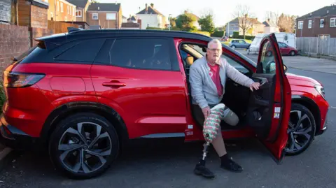 BBC News A man sits in the driver's seat of a large red car with the door open. The man is holding a prosthetic leg, and is wearing a grey zipped hoodie and shorts. 
