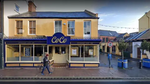 Cafe Coco photographed from across the street. It has been raining. A man with a bicycle is walking in front.