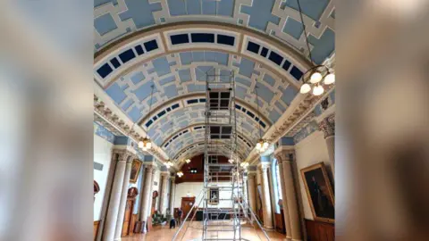 Colchester City Council Ornate pale blue and cream curved ceiling in room with pillars and oil paintings on wall. A scaffold stands in the centre of the room and reaches to the ceiling.