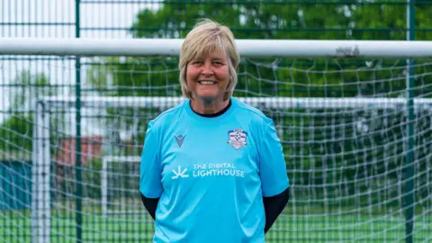 Simon Thurlow Janette Hynes wearing a blue football shirt in front of a goal on a football pitch. There are trees in the background.