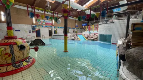 Jamie Niblock/BBC The swimming pool at Leisure World. The water is blue and runs over a white tiled floor. There are buckets that pour water over people suspended from a long yellow pole. There is a covered area with a waterfall in front of it. There are entrances to the changing rooms in the background and a blue slide.