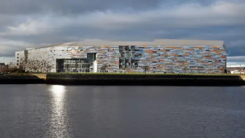 LDRS A general view of Middlesbrough College taken across the dock, with the water in the foreground. The large building is clad with grey, blue and orange material creating an irregular pattern.