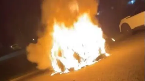 A large orange flame is seen coming from a parked car. A nearby white car is parked a few feet away.