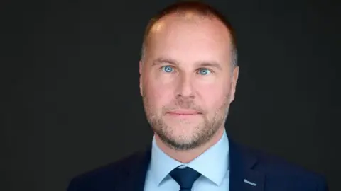 ASCL NI A man with short grey hair, wearing a navy suit, blue shirt and tie, pictured against a grey background