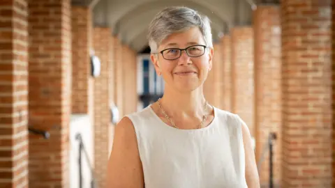 A woman with short grey hair and glasses. In the background is a brick walkway. 