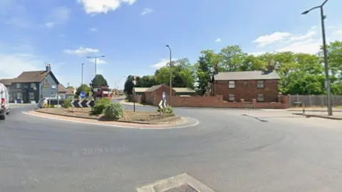 A suburban roundabout pictured in daylight. A brick house can be seen at one side of the roundabout and a pub on the other. The roundabout itself is oblong shape and has small bushes on it.