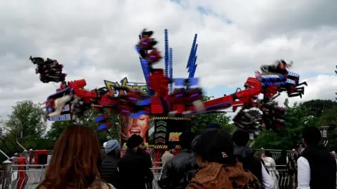 A red fairground ride in motion, with the ride slightly blurred because of the movement. A queue of people can be seen in front of the ride.