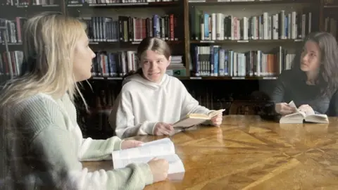 Three female students sitting at a table with books with shelves full of books behind them 