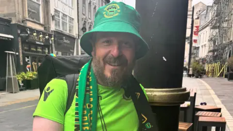 Dave Chapman is wearing a green T-shirt and green bucket hat and is standing outside. He is looking directly at the camera and smiling. He has a brown beard. 