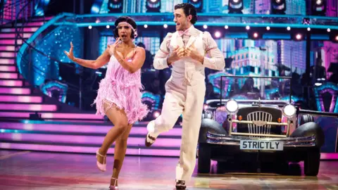 Balvinder Sopal in 1920s flapper dress with her dance partner Julian Caillon in a white suit dancing on stage at Strictly with a vintage car behind them