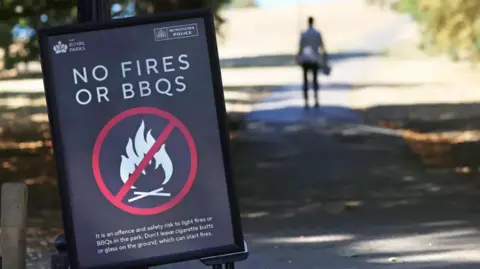EPA A 'No fires or BBQs' sign in a park, with a person walking down a path, silhouetted in the background