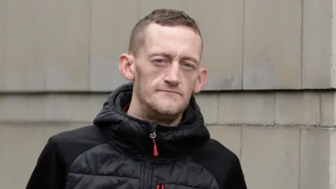 Craig Rowland - a man looks at the camera as he stands outside a white-stone building. He has short brown hair and is wearing a black puffed jacket with a red zip.