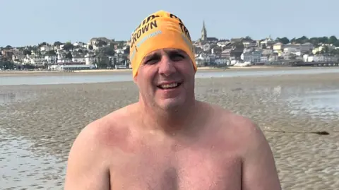 Simon, sitting on the beach with his shirt off after completing the swim