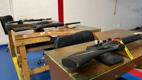 Tom Jackson/BBC Three rifles have been lay on three wooden tables in a white bricked room.