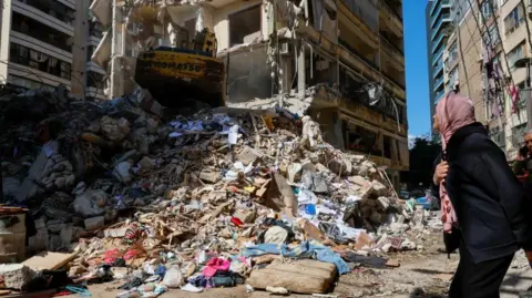 A woman wearing black and with a pale red headscarf looks at a bulldozer clearing debris and rubble at a building hit by a strike