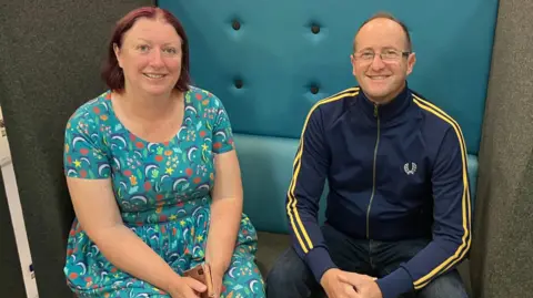 Joanne Kerr on the left wearing a dress smiling at the camera and Will Davies on the right wearing a blue jacket and glasses smiling at the camera, sitting in a grey and blue booth chair.
