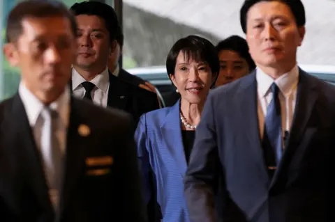 Reuters Japan's new Prime Minister Sanae Takaichi arrives at the prime minister's official residence in Tokyo, Japan October 21, 2025. She is wearing a blue suit, with a pearl necklace and earings. Her hair is short. She is surrounded on all sides by men in suits