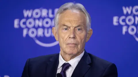 La EPA Sir Tony Blair con traje de marina y corbata sentada frente a una bandera azul en el Foro Económico Mundial.