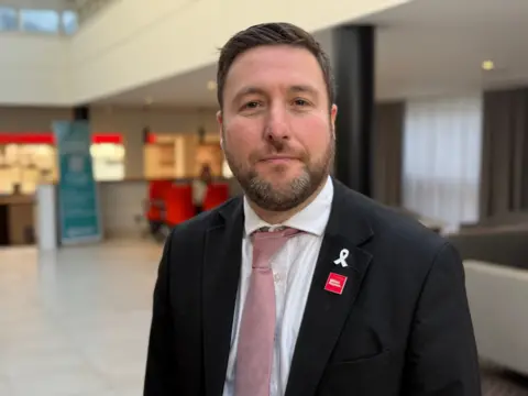 Sam Read/BBC A picture of a man who has short dark brown hair and a dark brown beard with white bits. He is wearing a black suit jacket with a dark pink tie.