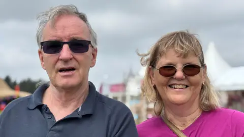 A man with grey hair and black sunglasses is wearing a navy t-shirt. A woman stands next to him wearing a pink top. She has shoulder-length blonde hair and tinted glasses.