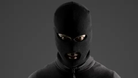 A person stares at the camera while wearing a full-face balaclava and a zipped up, grey jumper. They are standing in front of a dark grey wall.
