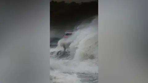 Paul Moane Strong winds throw waves over a white car that has red break lights.