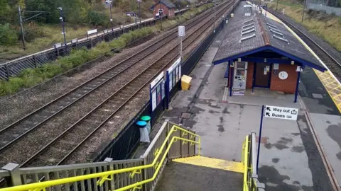 The image shows a railway station viewed from the top of a footbridge. In the foreground, bright yellow handrails lead down a set of stairs toward the platforms. To the right is a platform with a small brick station building that has a blue roof and several skylights. Signs point to the “Way out” and “Buses.” Multiple railway tracks run through the center of the scene. On the left side is another platform.